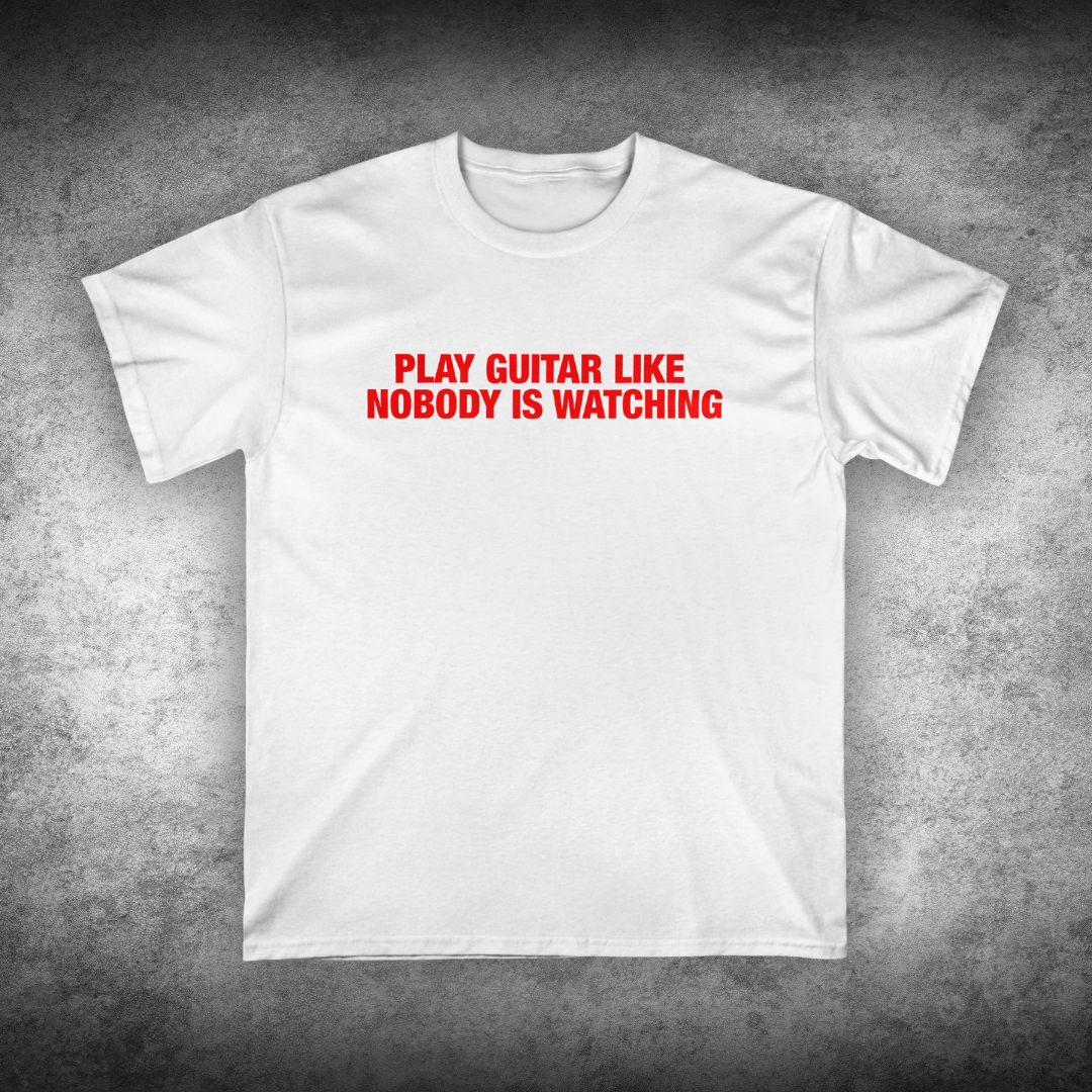 White t-shirt with red text 'Play Guitar Like Nobody is Watching' on a textured gray background