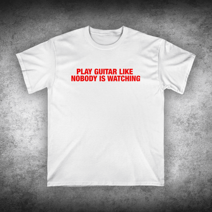 White t-shirt with red text 'Play Guitar Like Nobody is Watching' on a textured gray background