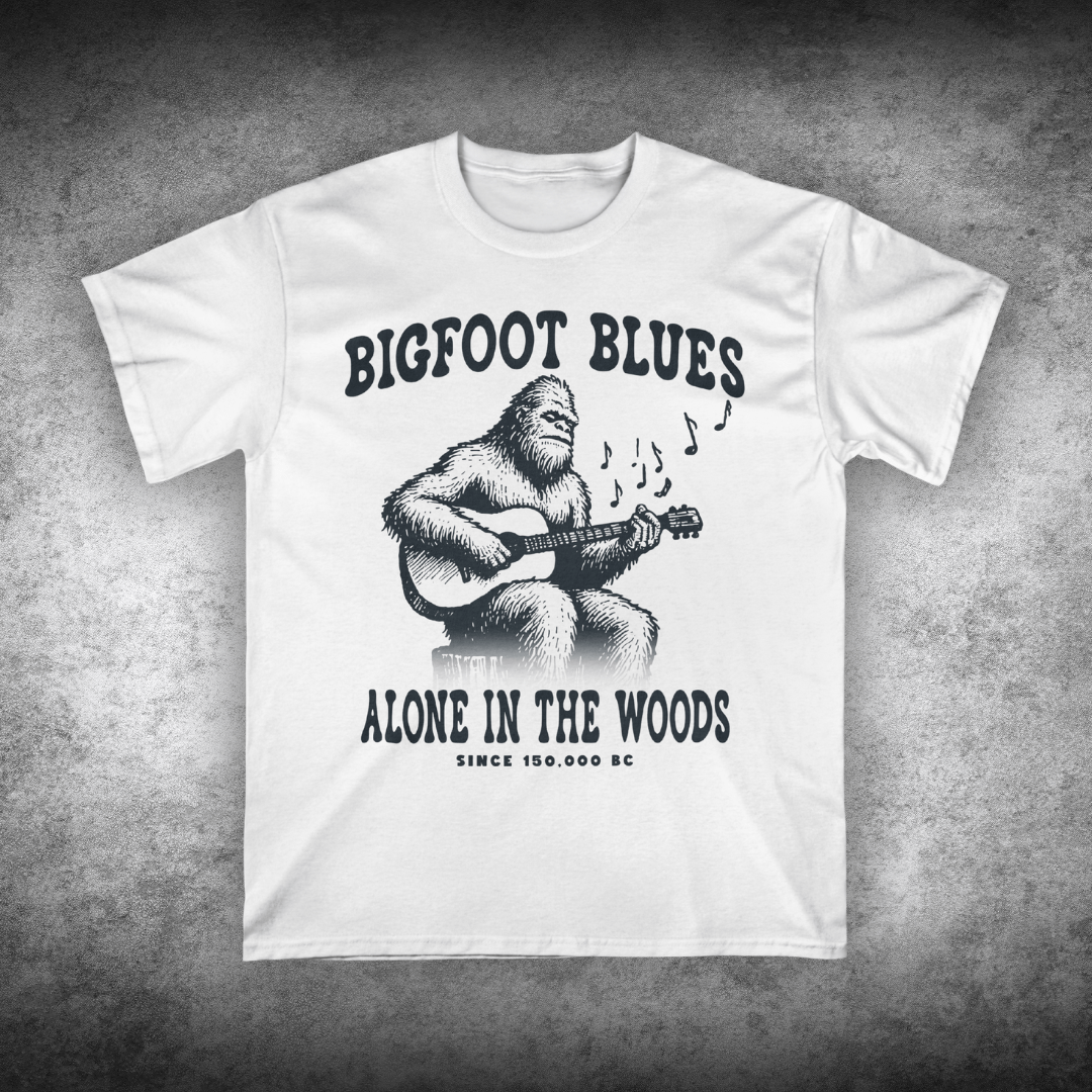 White t-shirt with a graphic of a bigfoot playing guitar and text on a textured gray background