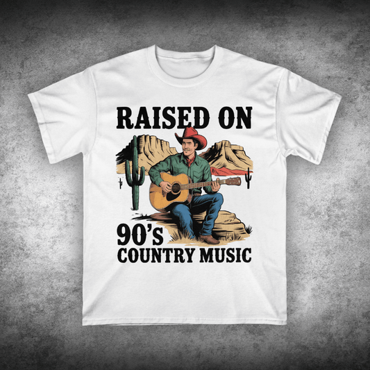 White t-shirt with a graphic of a cowboy playing guitar and text 'Raised on 90's Country Music' on a textured gray background.